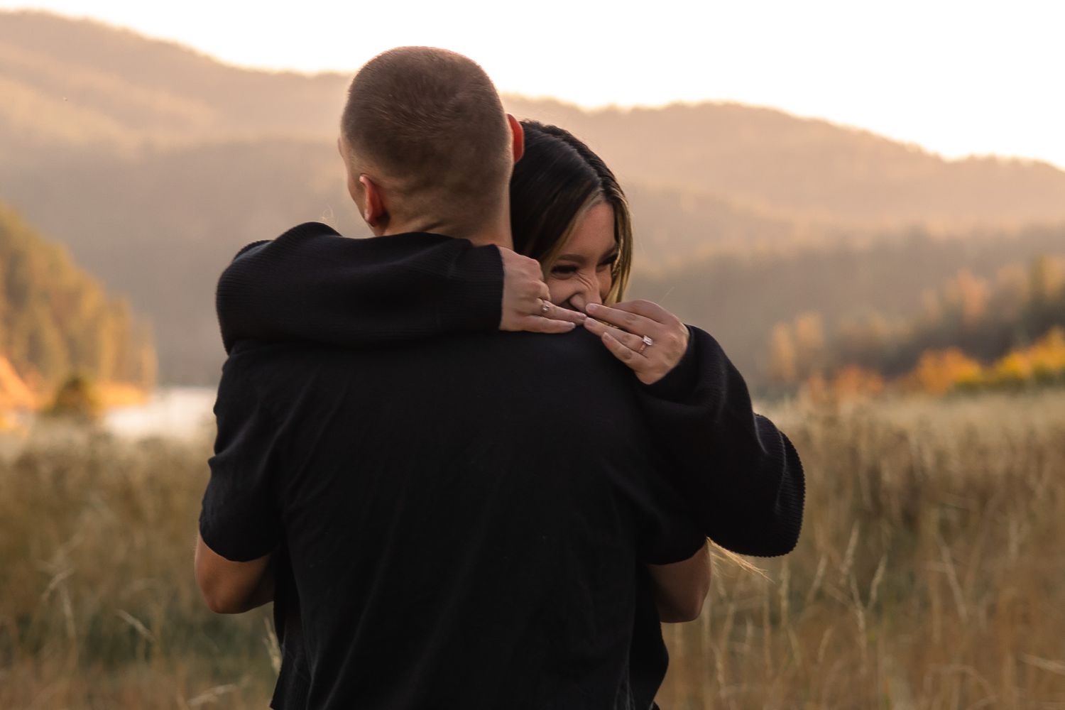 Couple embraces while watching sunset in golden field with mountains in background.