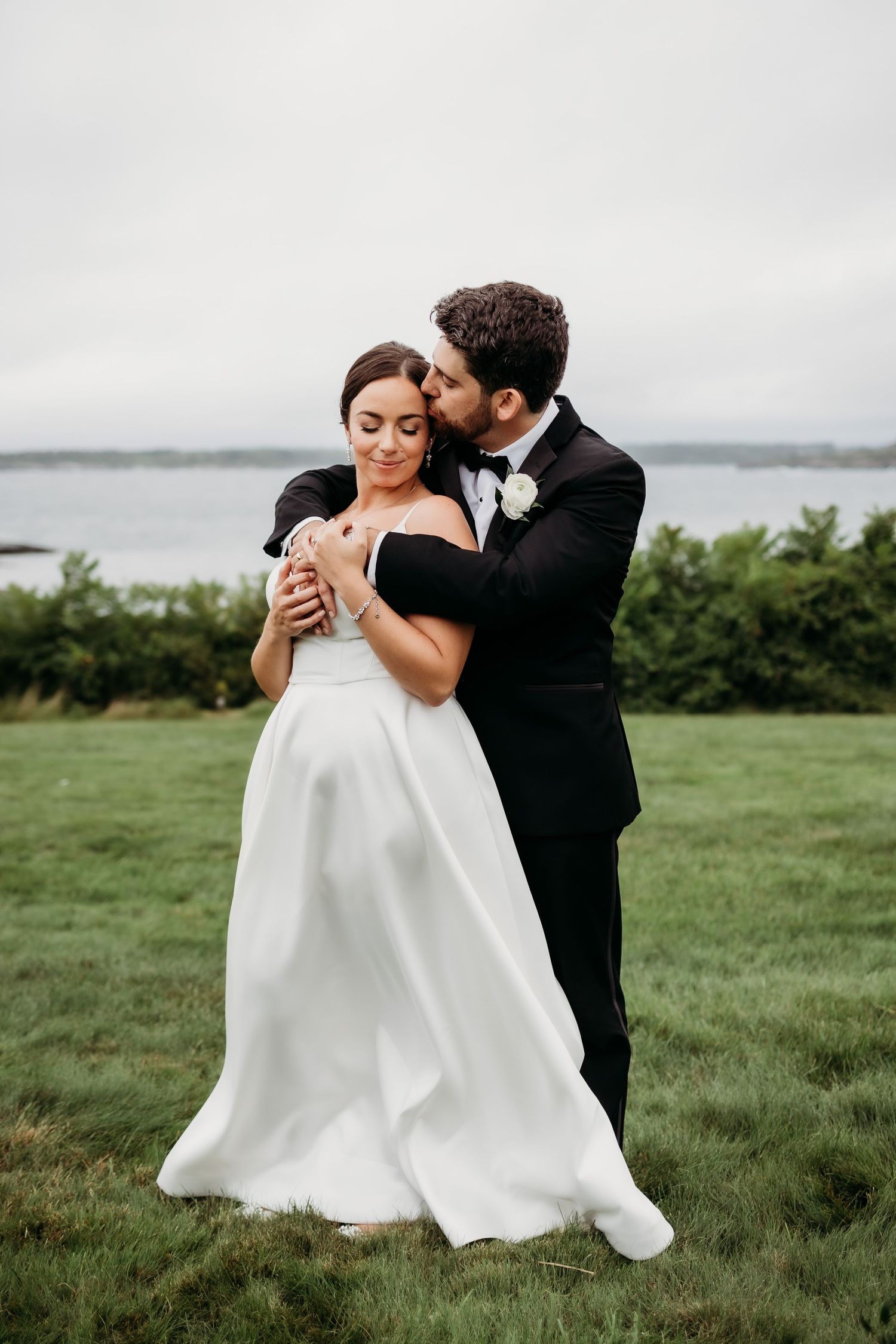 Romantic wedding couple embracing in windswept field with overcast sky.