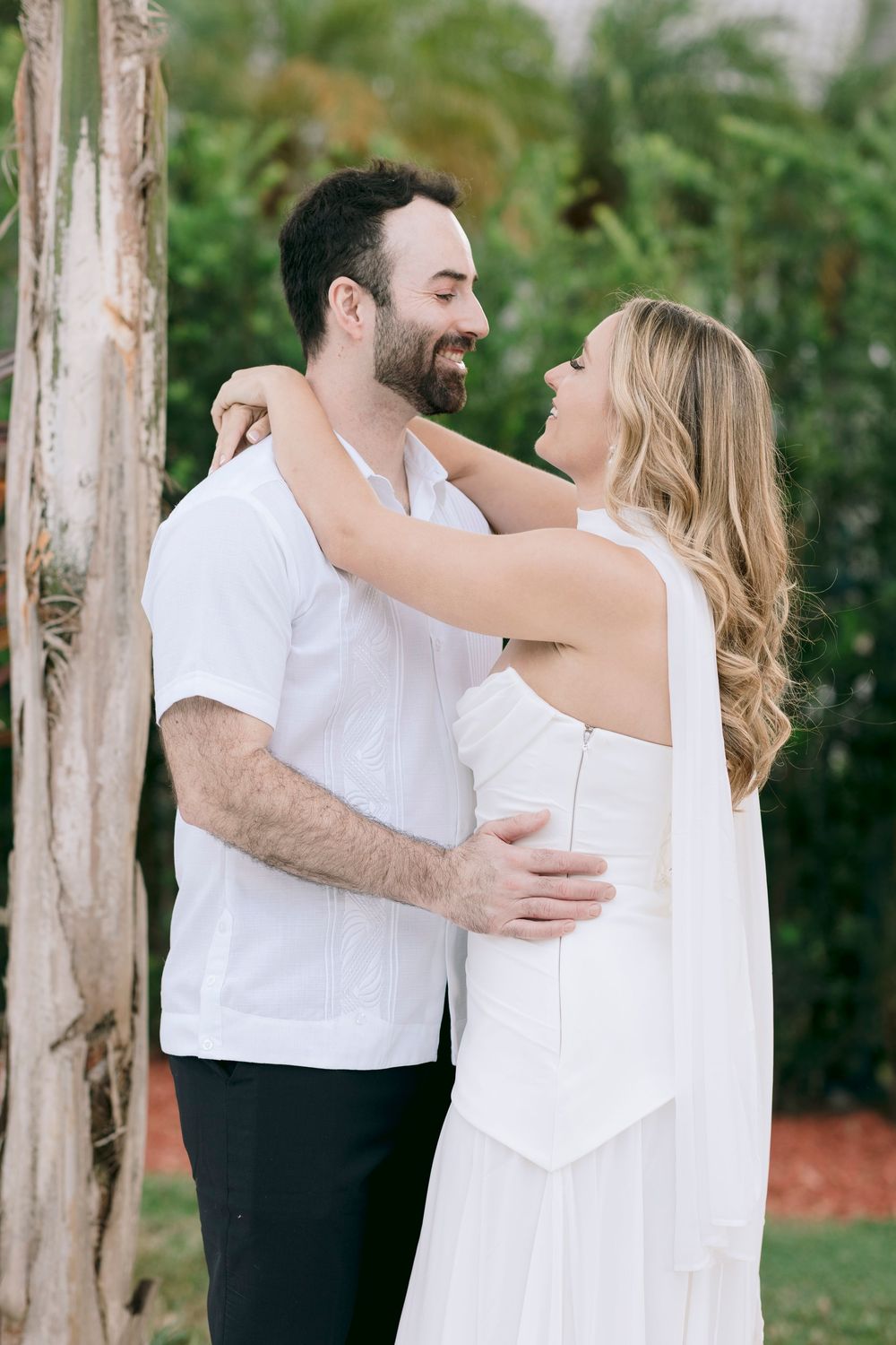 A romantic couple in white outfits embrace and laugh together outdoors against a lush green garden backdrop.