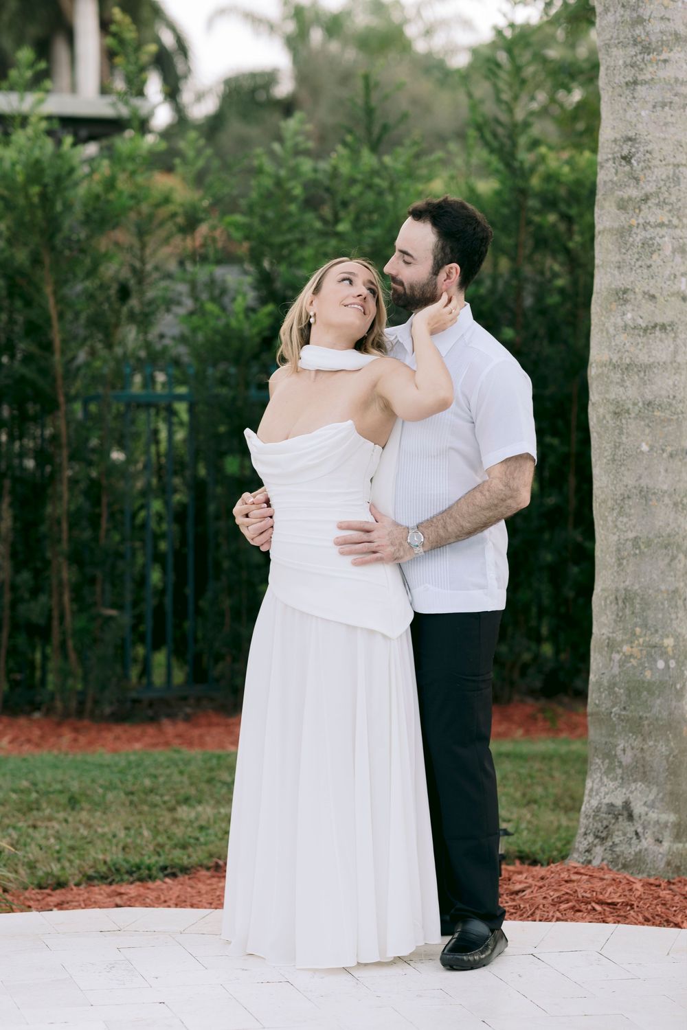 A romantic couple in white and black attire shares an intimate dance moment under palm trees in a garden setting.