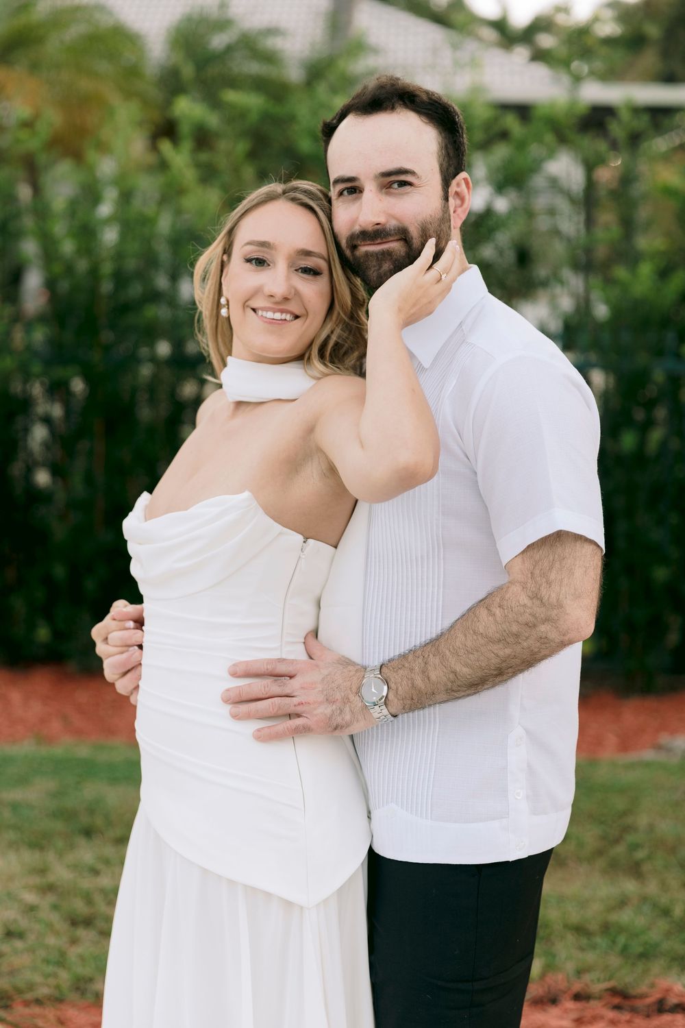 A couple in wedding attire embraces in a romantic outdoor photoshoot with greenery in the background.
