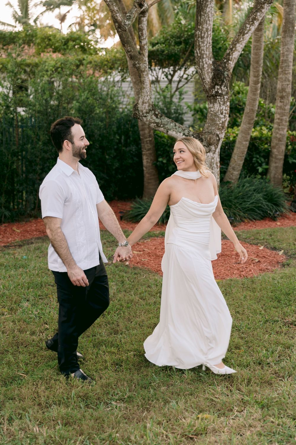A couple in wedding attire dance and twirl playfully in a sunlit garden setting with trees in the background.