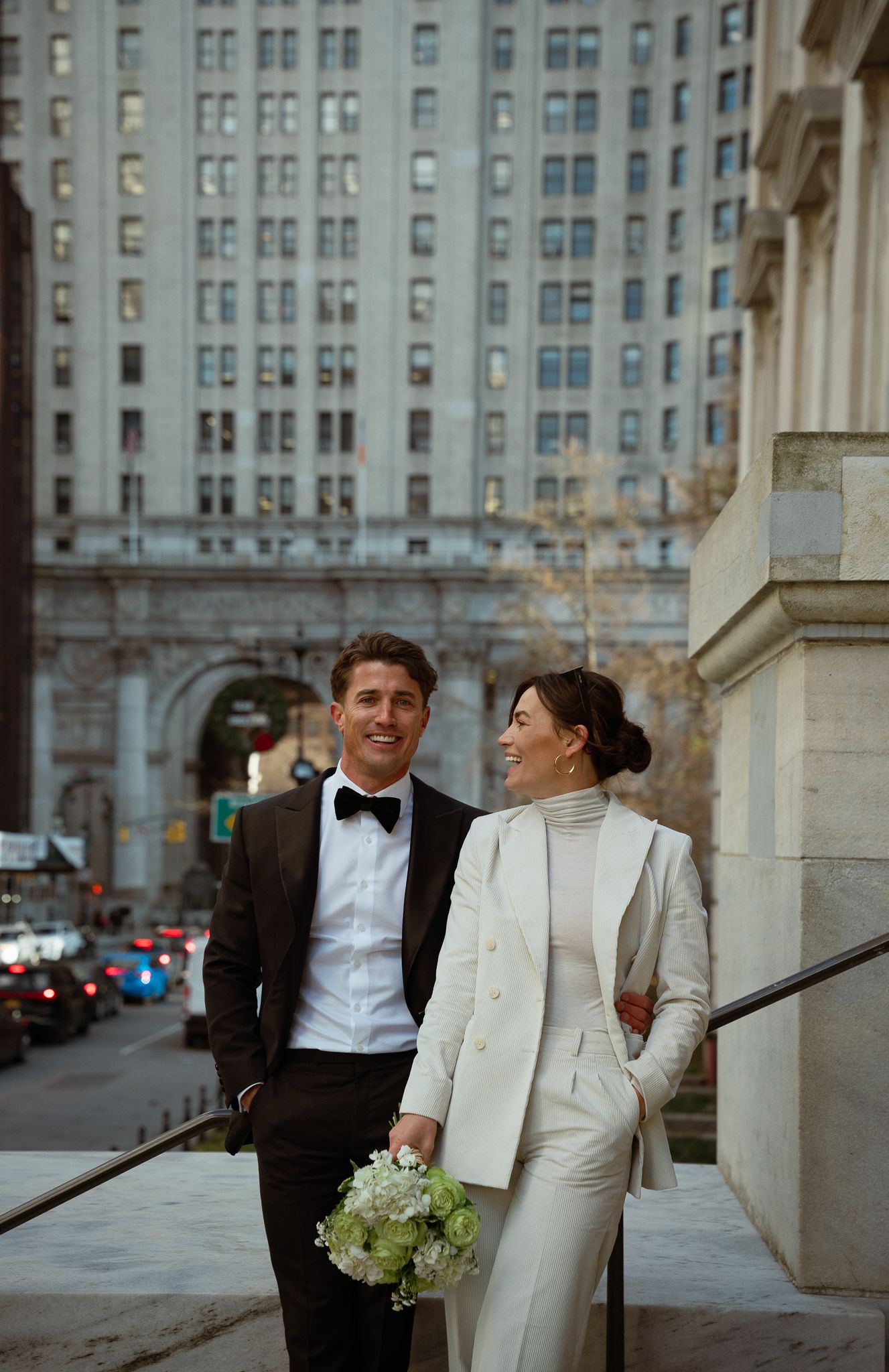 A couple shares a tender moment outside a grand building with urban architecture behind them.