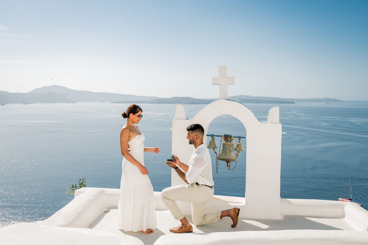 A romantic wedding proposal takes place on a white terrace overlooking the blue Mediterranean Sea in Santorini, Greece.