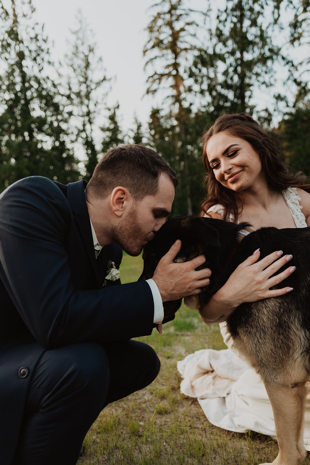 Bride and groom in wedding attire kneel to kiss their German Shepherd dog in a forest setting.