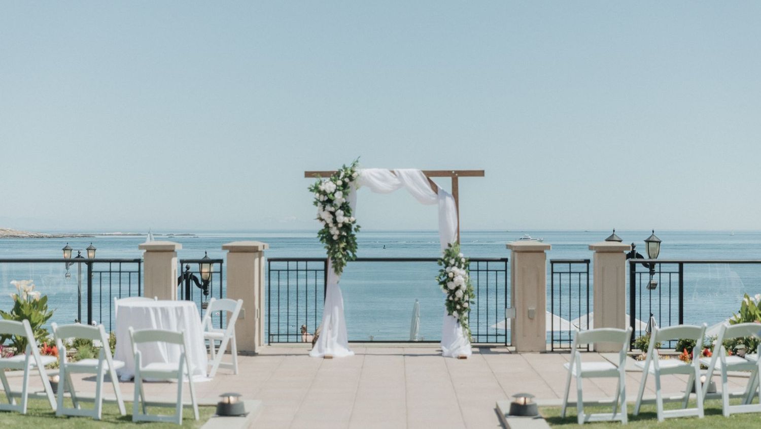 Elegant outdoor wedding ceremony setup with floral arch, white chairs, and stunning ocean view under clear blue sky.