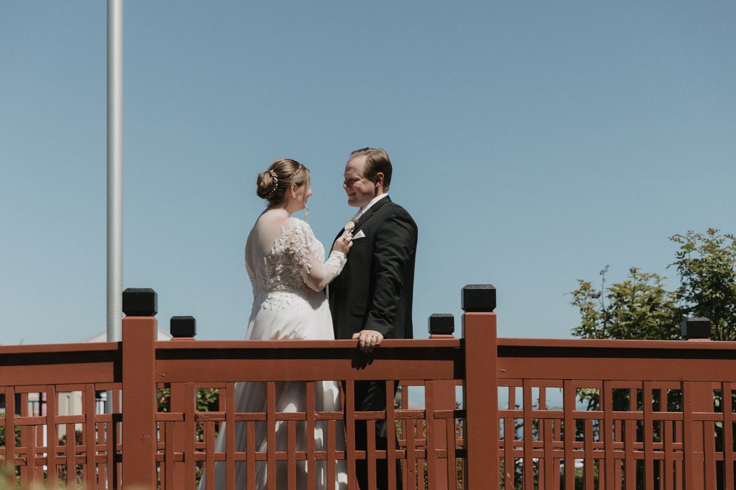 Bride and groom share a tender moment on a red wooden bridge with clear blue sky and ocean in the background.