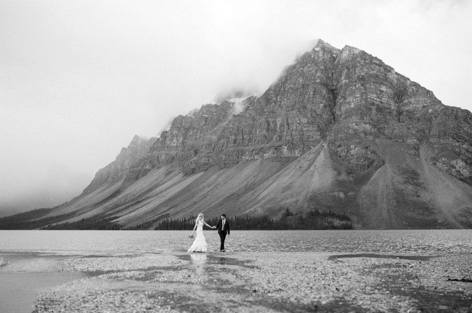 bride and groom hold hands and walk along the edge of a mountain lake in Kananaskis