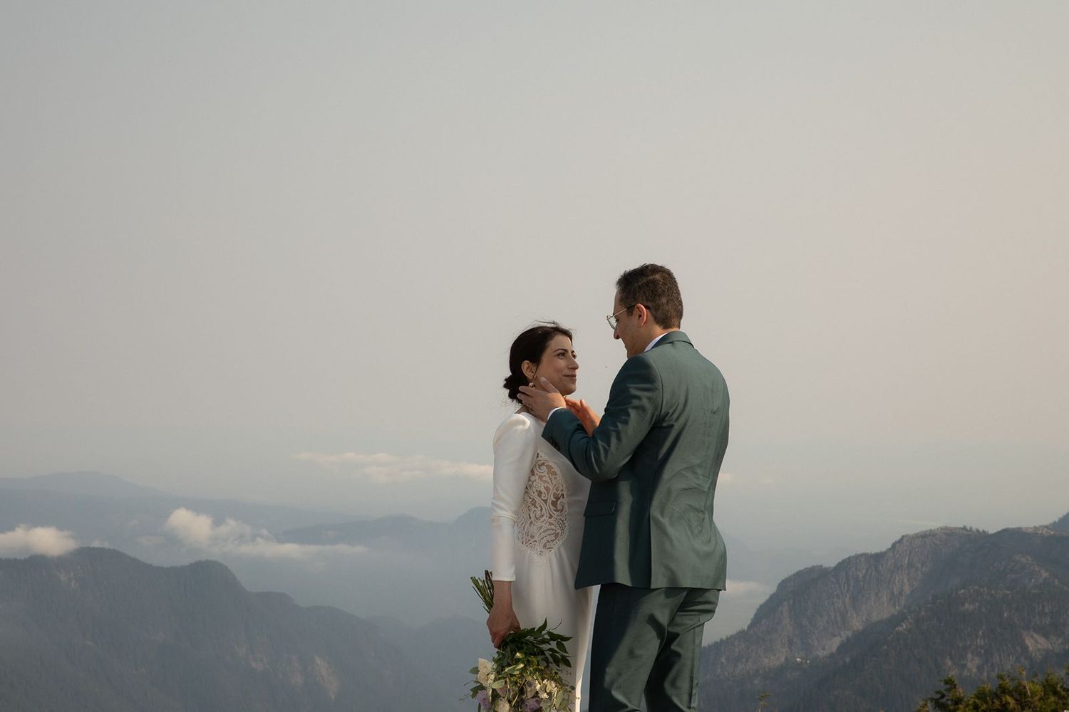 Groom tenderly touches brides face while holding bouquet with misty mountain valley in background.