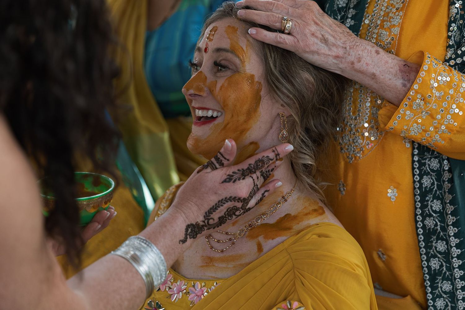Smiling woman having turmeric haldi paste applied to her face and neck at Indian wedding ceremony.