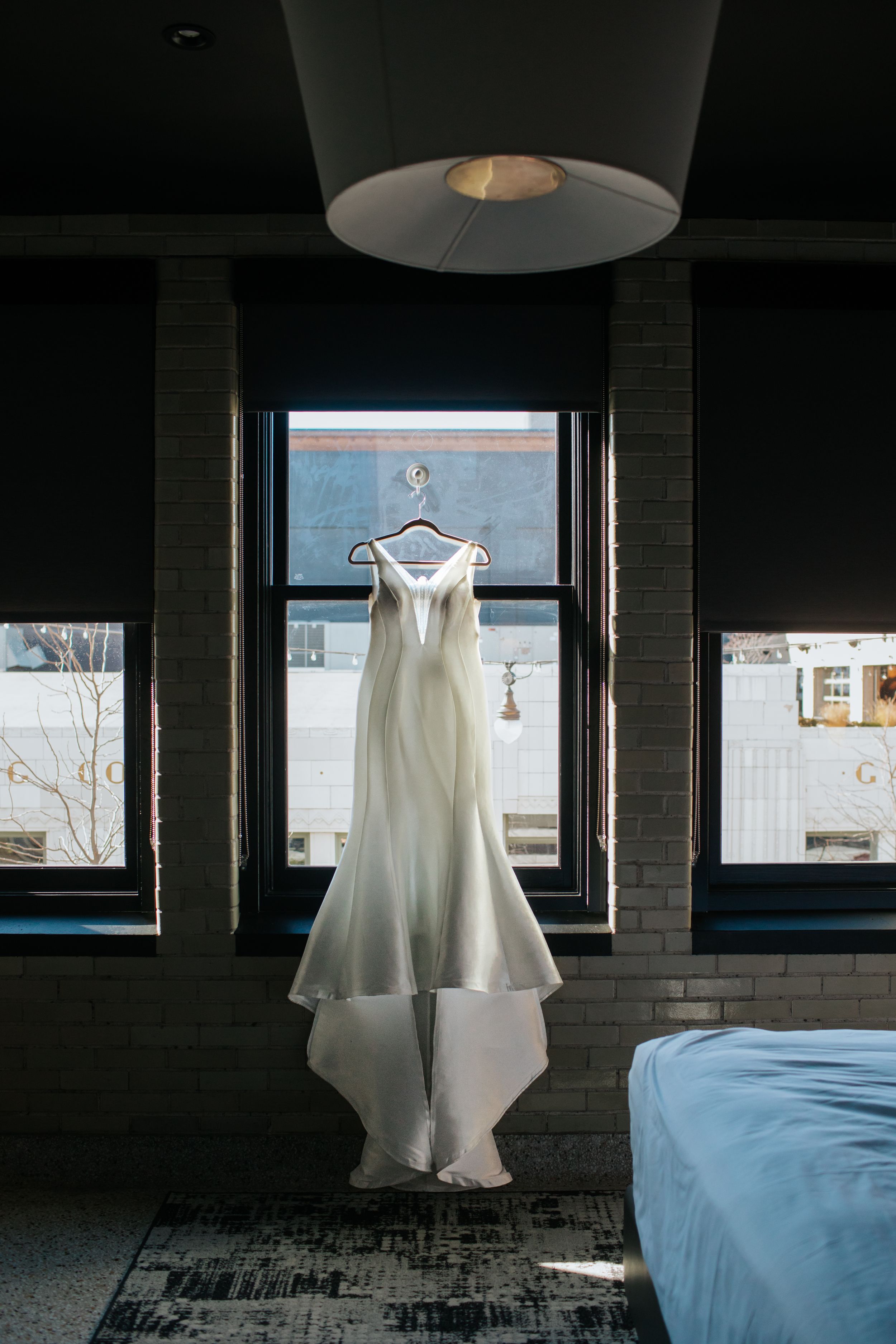 A white wedding dress hangs in a dark room with natural light streaming through windows, creating a serene atmosphere.