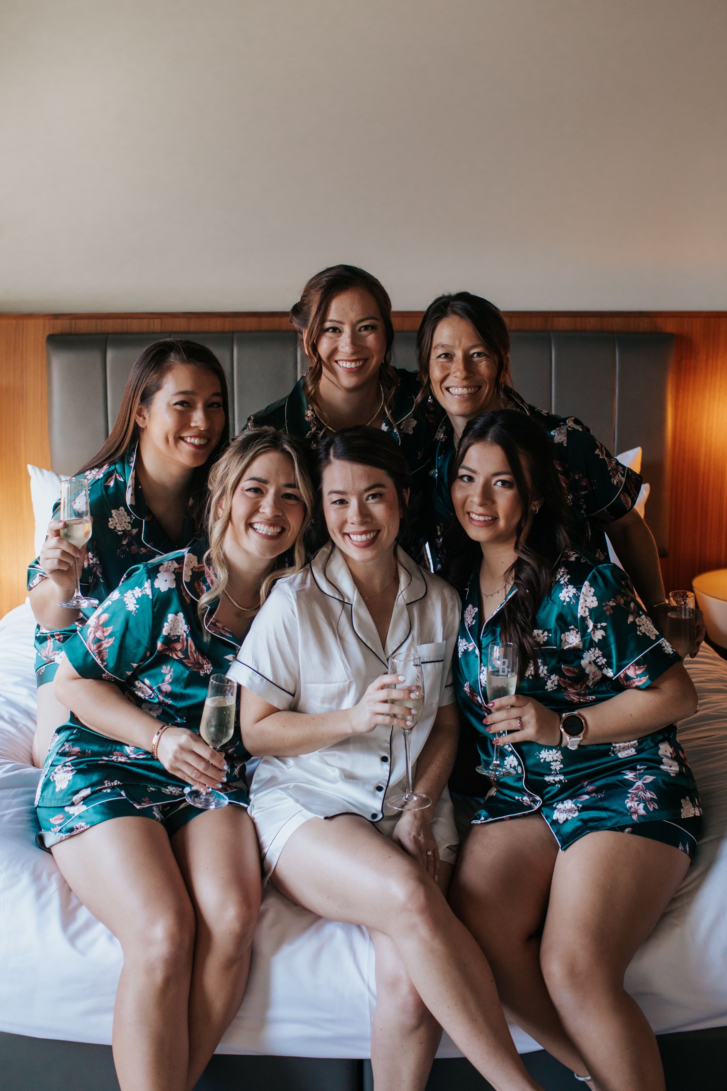 A group wearing matching floral robes lounges together on a white bed in a hotel room during a celebration.