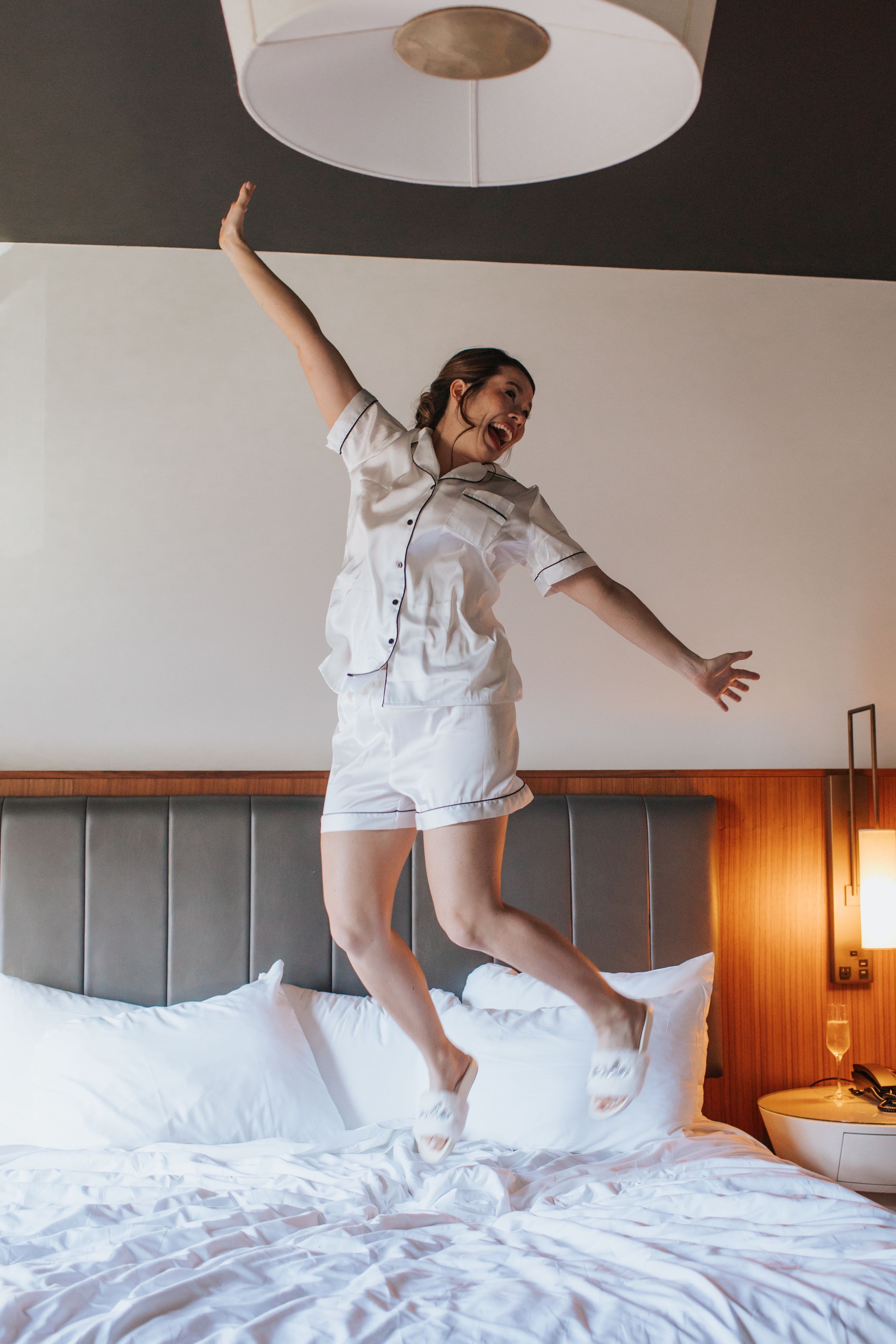 Person in white outfit jumping playfully on a hotel bed with white linens and wooden headboard.