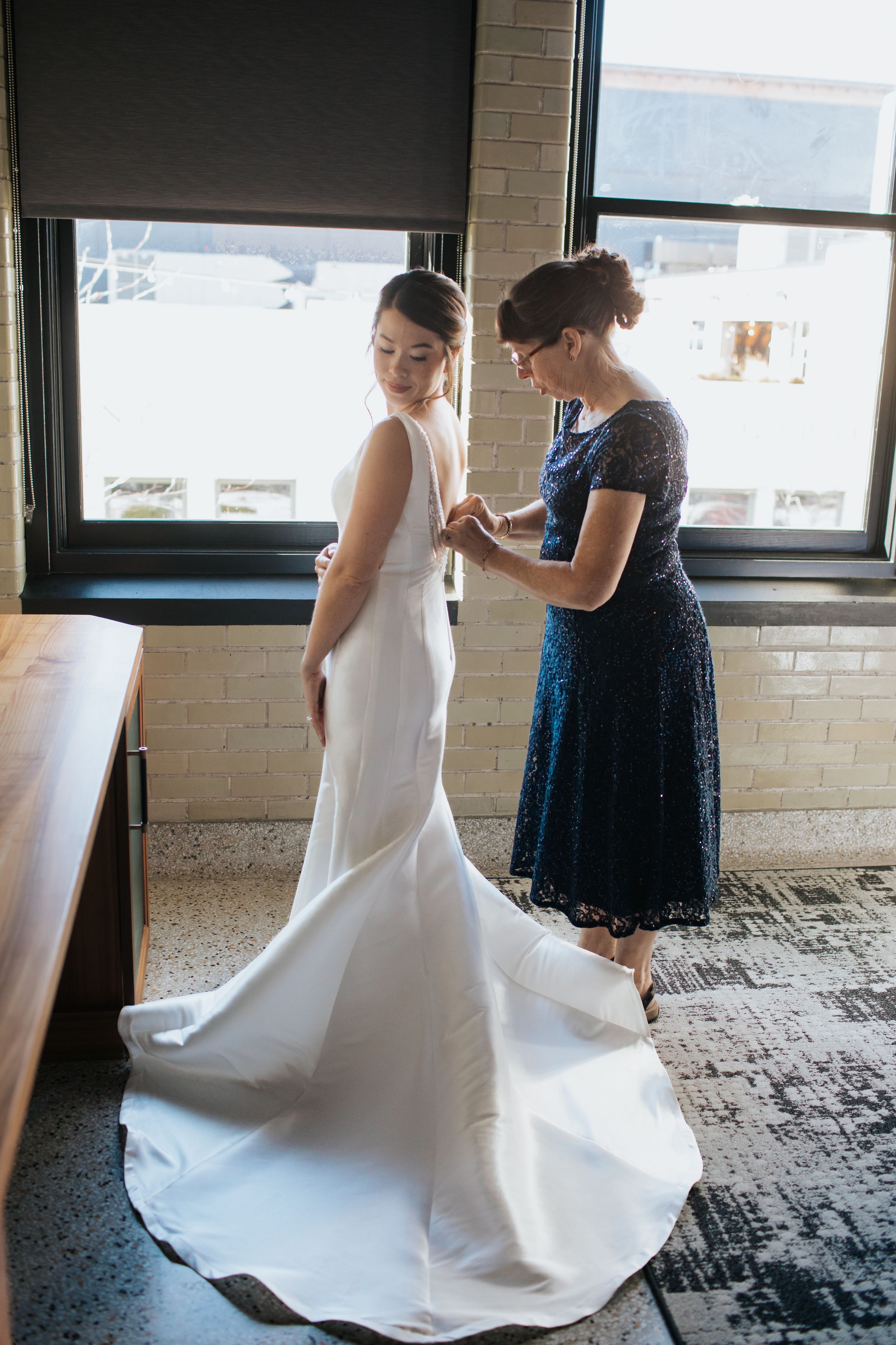Wedding preparation scene showing a white bridal gown being adjusted in a room with brick walls and large windows.