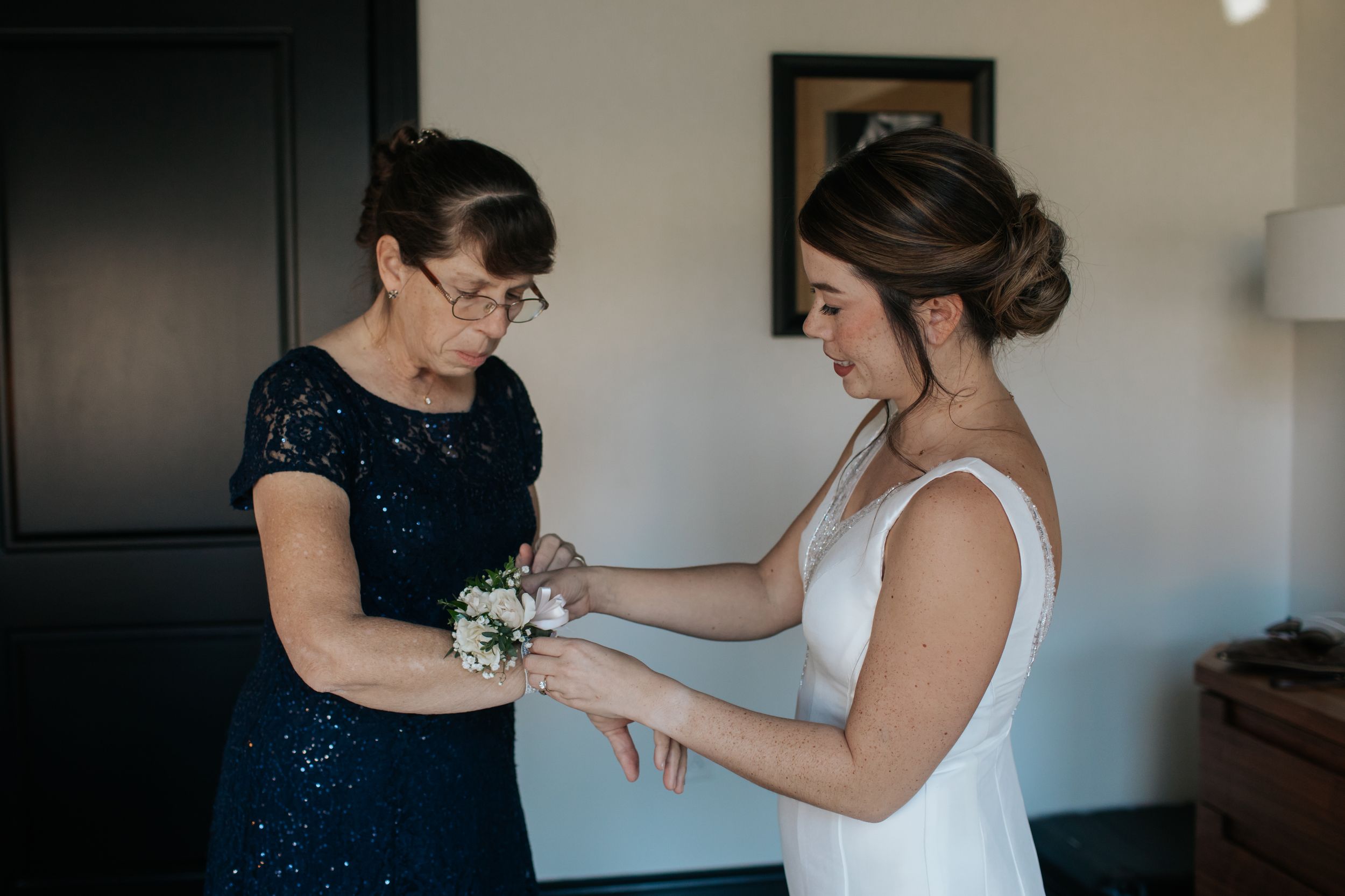 A woman in a white wedding dress gets help with her corsage from someone wearing a navy blue dress.