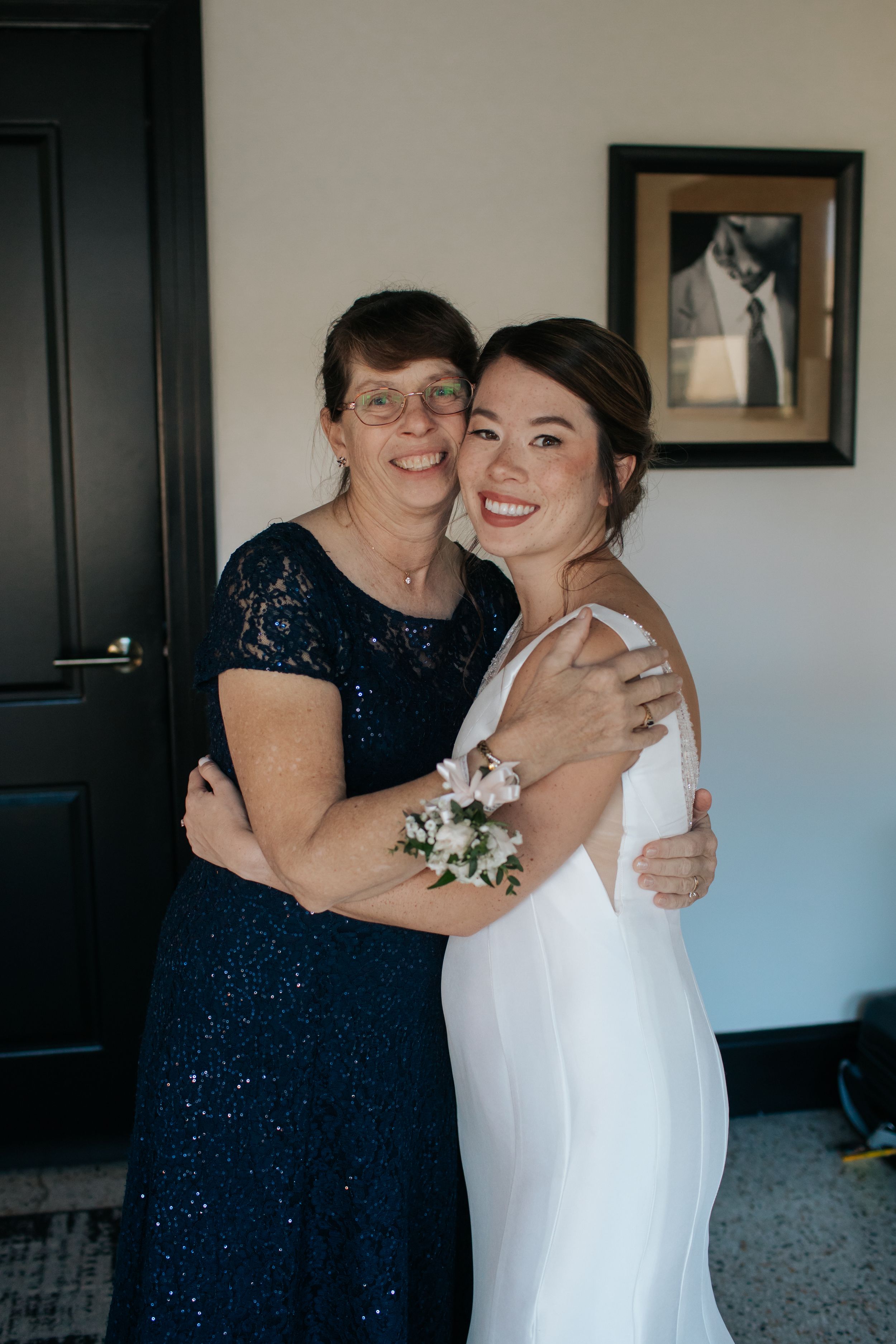Two women embrace in formal attire at a wedding, one wearing a navy lace dress and one in a white bridal gown.