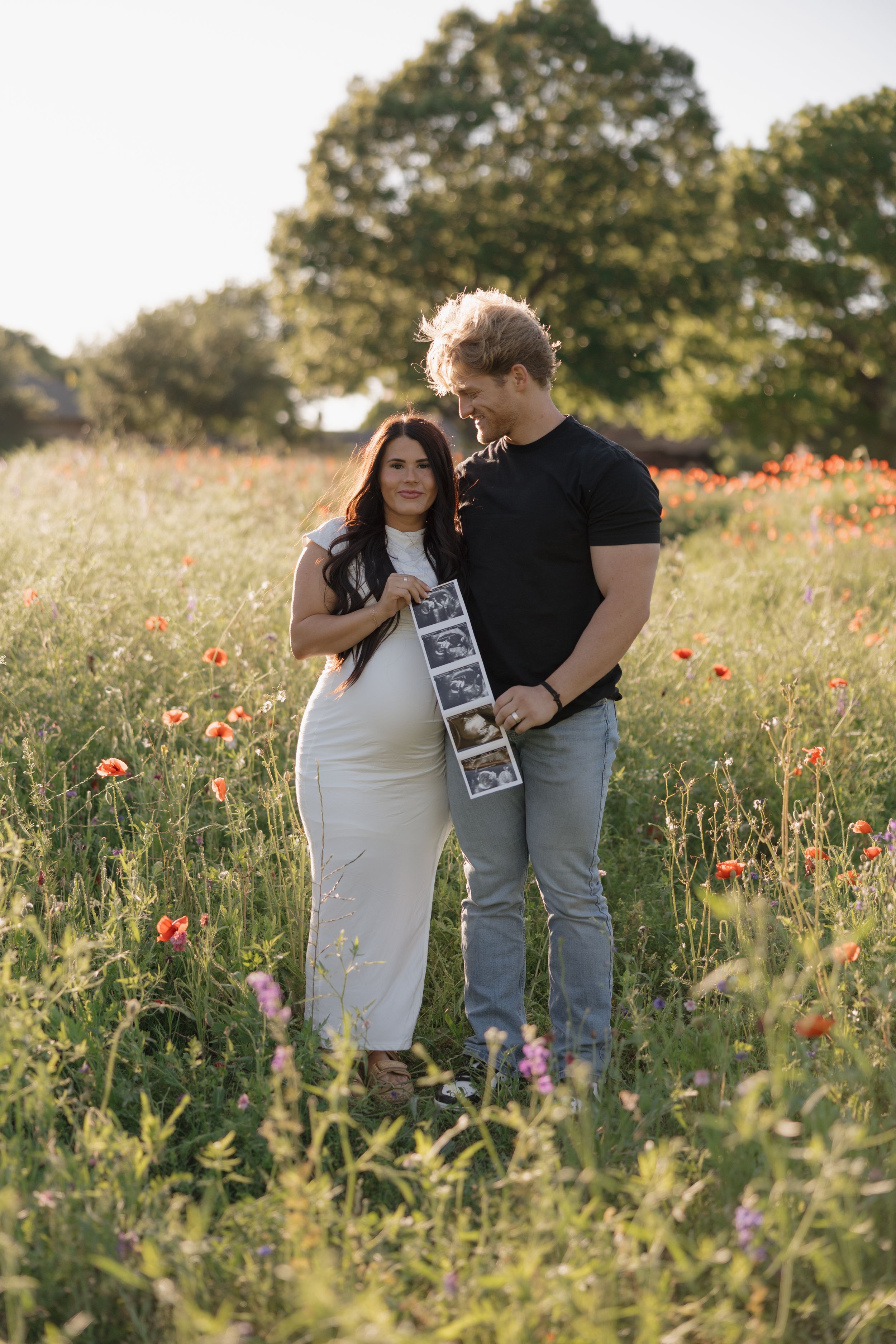A couple stands together in a wildflower meadow at sunset holding ultrasound photos in a sweet pregnancy announcement.