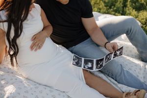 Couple holding ultrasound photos while sitting together on white blanket outdoors in sunlight.