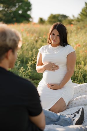 Pregnant woman in white dress sitting on blanket in field of wildflowers during sunset photoshoot.