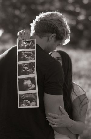Black and white photo of a couple embracing while looking at ultrasound images in a romantic outdoor setting.