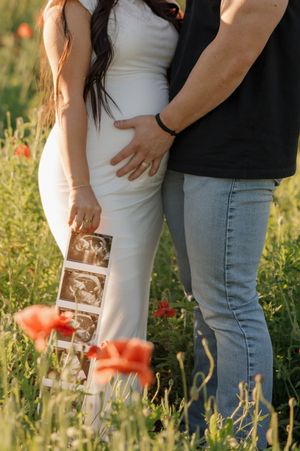 A couple stands in a wildflower field holding ultrasound photos while wearing white dress and casual attire with poppies nearby.