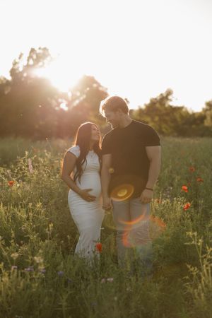 A romantic couple stands together in a wildflower meadow with poppies at golden hour sunset.