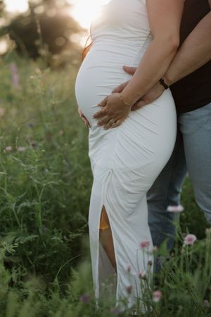 A romantic maternity photo showing a couple embracing in a wildflower meadow at sunset, with the mother in a white dress.