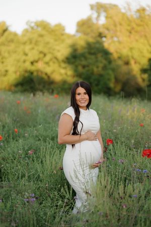 A pregnant woman in a white lace dress stands in a wildflower meadow at sunset with red poppies in bloom.