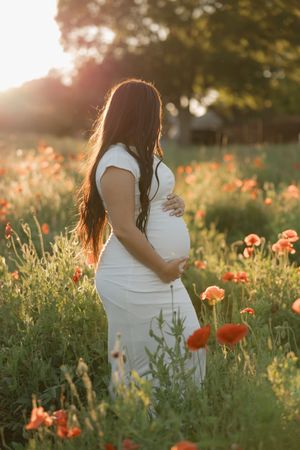 Expectant mother in white dress standing among vibrant red poppies at golden sunset in summer field.