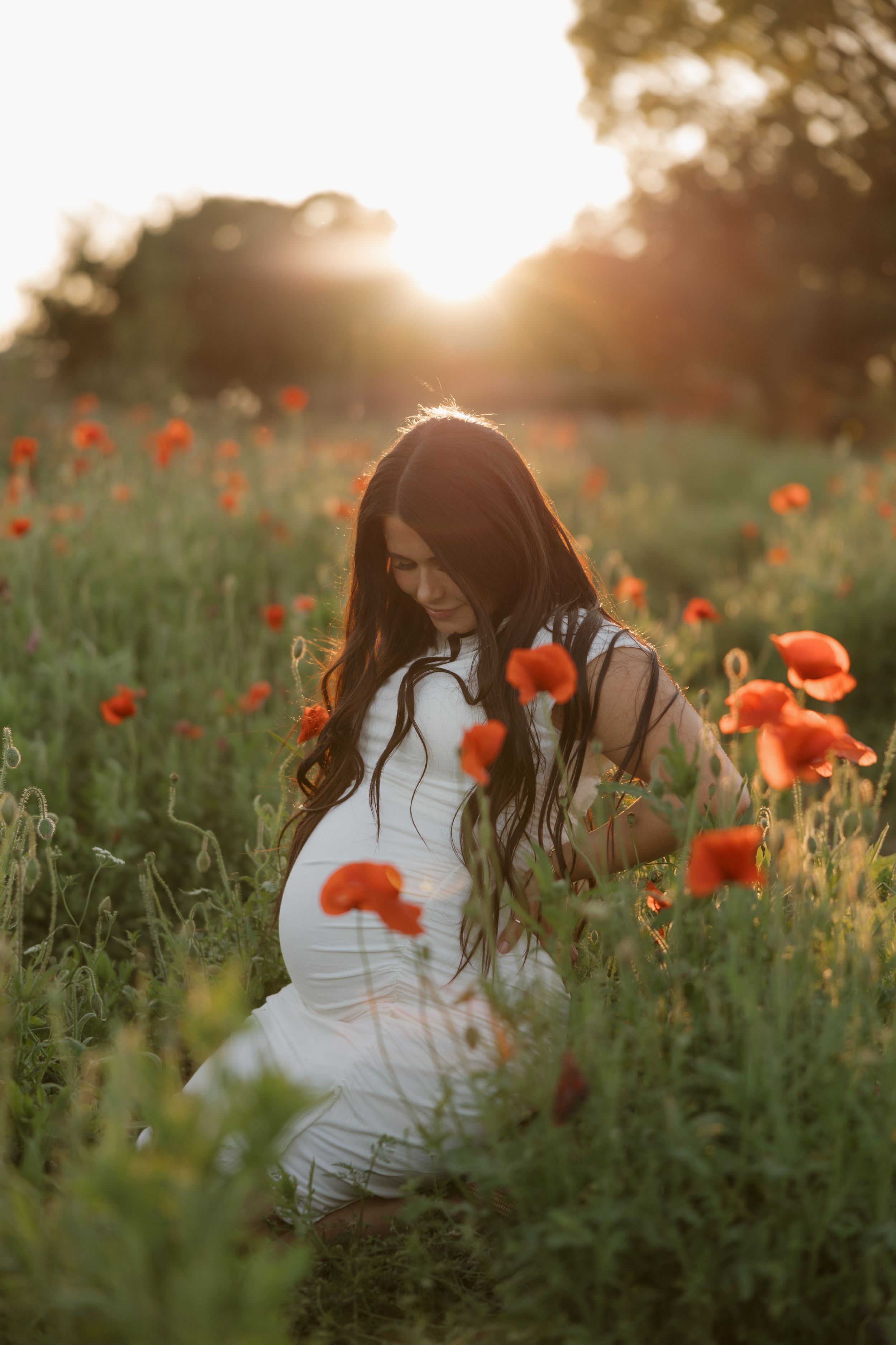 Silhouette of woman in white dress standing in field of red poppies at golden sunset.