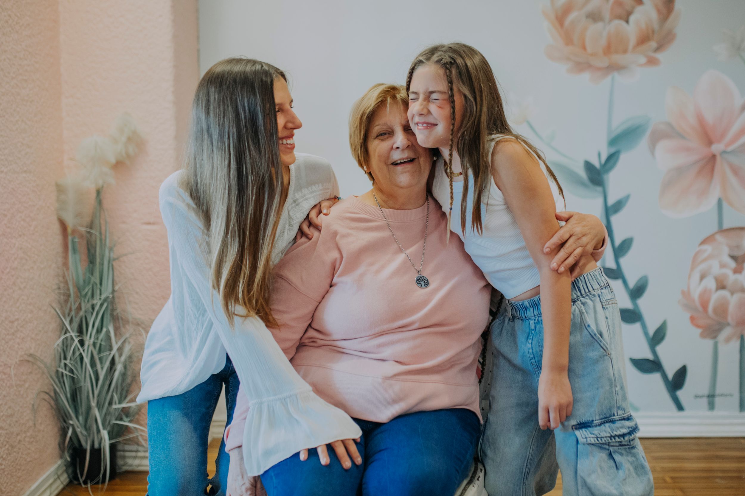 Three women sitting together and laughing against a floral wall backdrop in casual clothing.