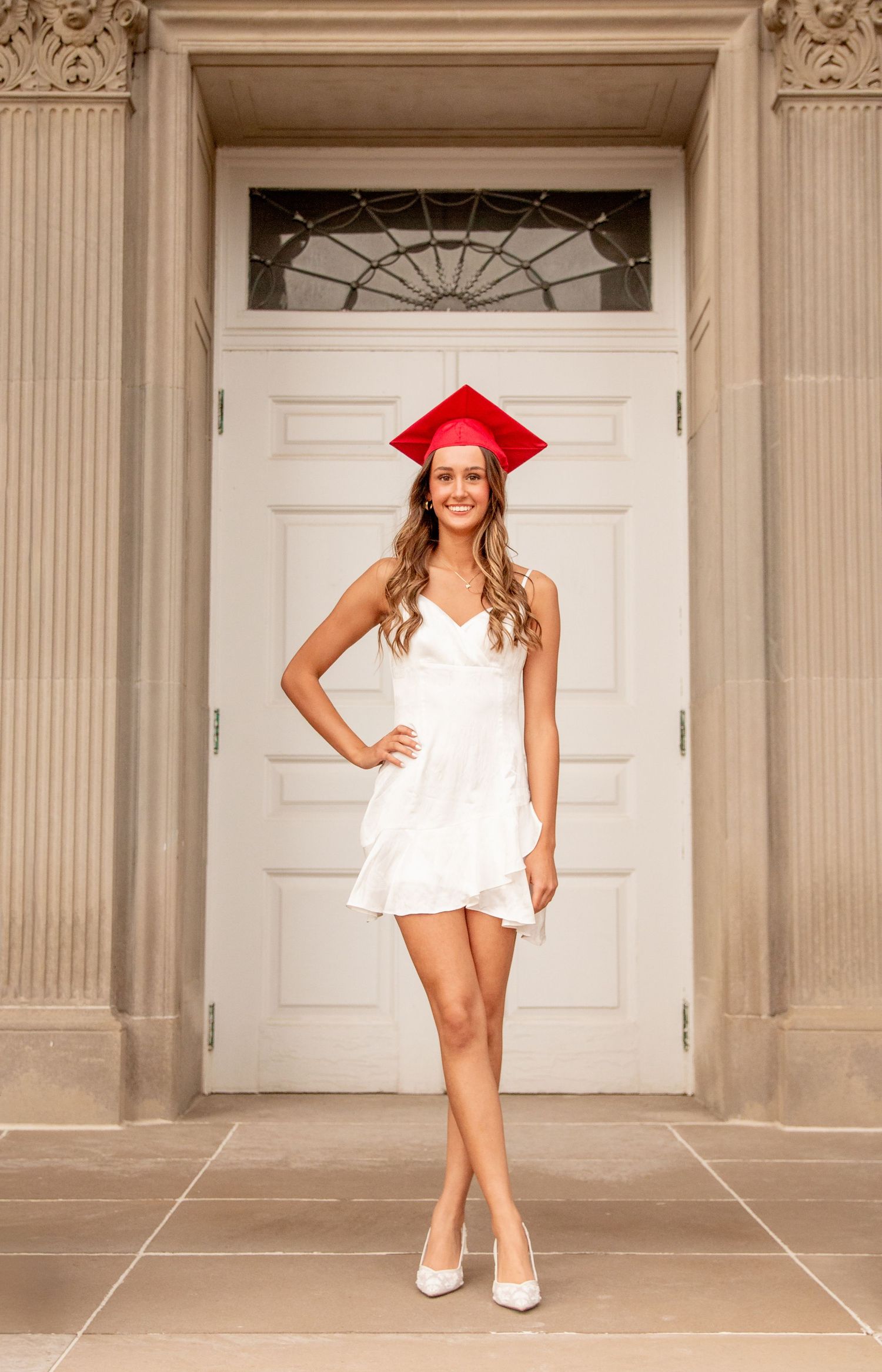 Smiling graduate in white dress and red cap posing before elegant white door.
