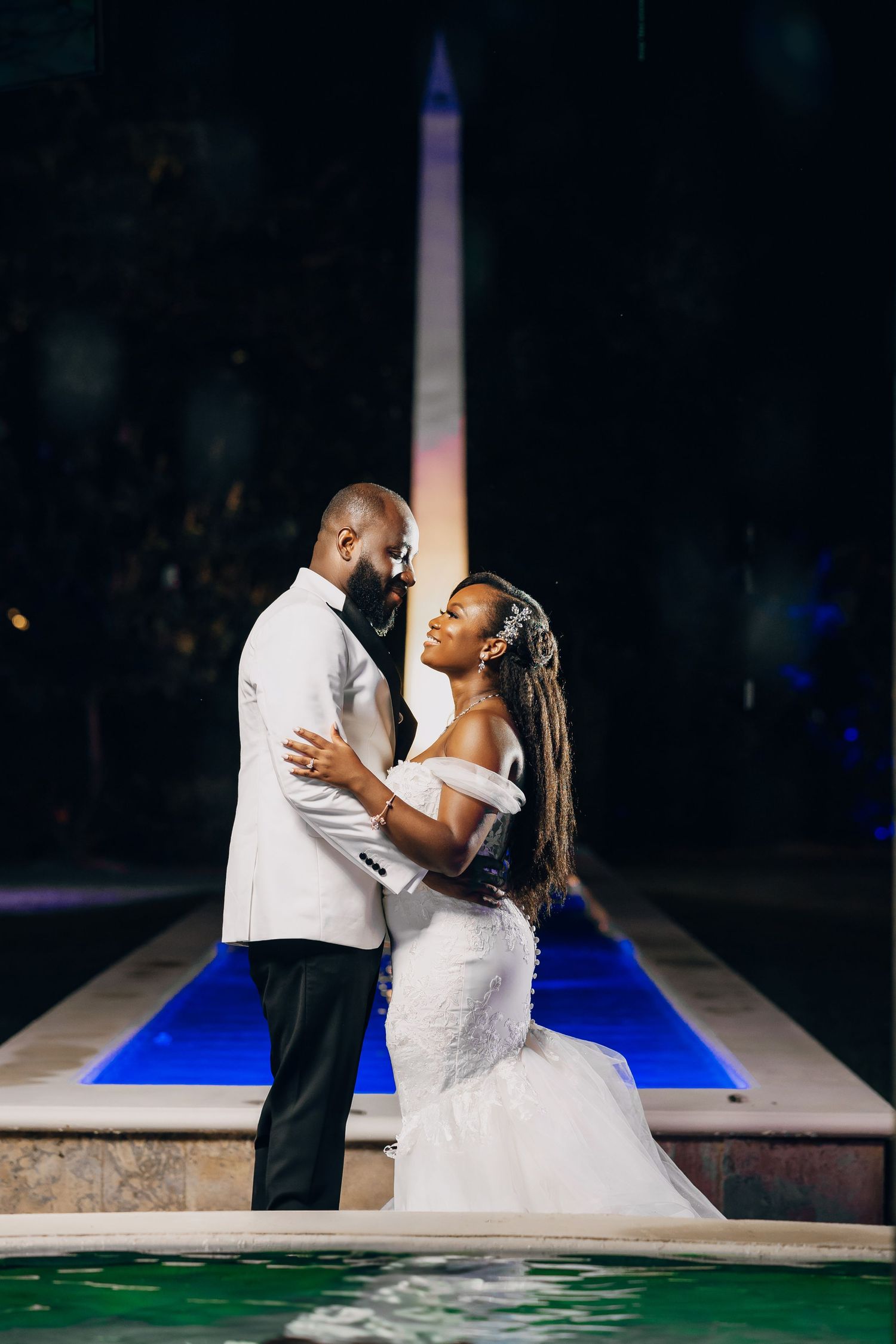 A couple shares a romantic moment in elegant wedding attire near a reflecting pool with the Washington Monument in view.