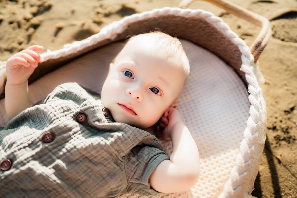 Baby in light gray clothing lying on blanket outdoors in bright sunlight on sandy ground.