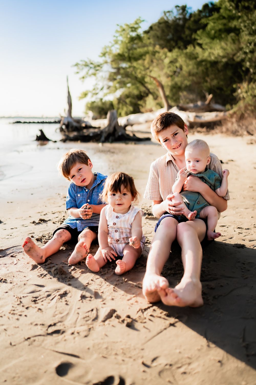 Family sitting together on a sandy beach with driftwood and trees in the background on a sunny day.