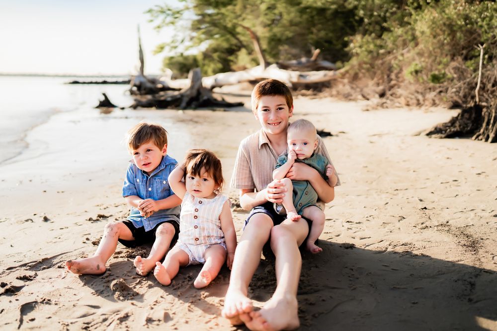 Family sits together on sandy beach near driftwood and water on sunny day while wearing casual summer attire.