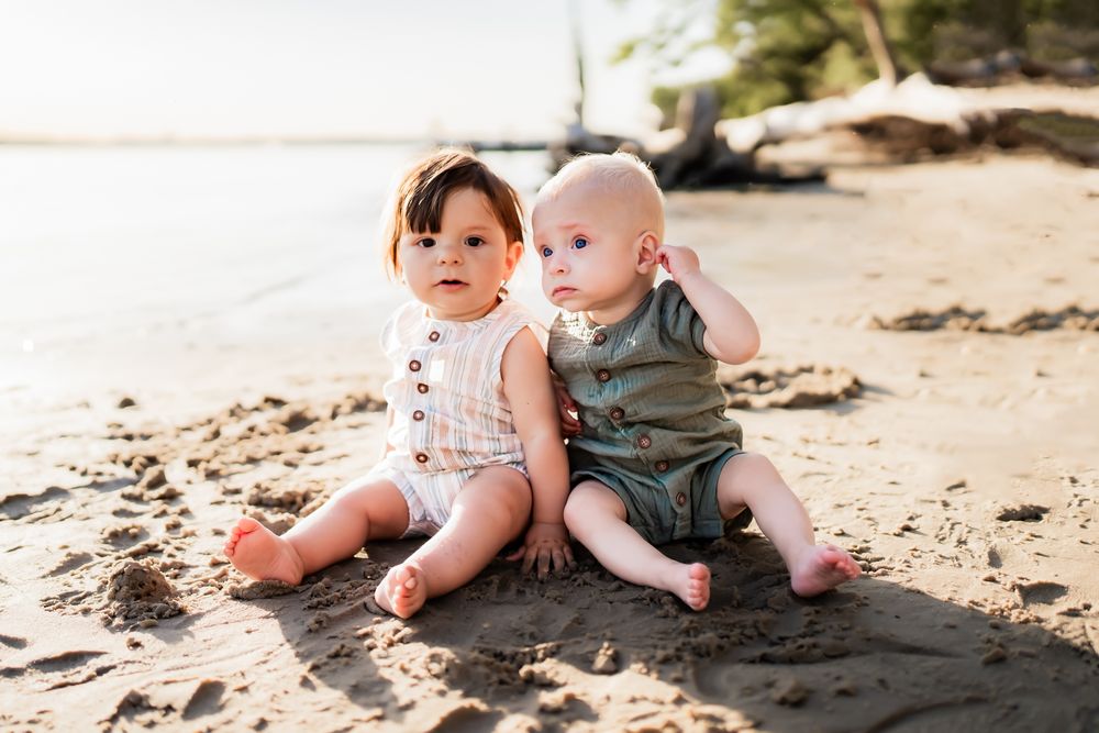 Two young toddlers in summer outfits sit together on a sunny beach with driftwood and sand in the background.