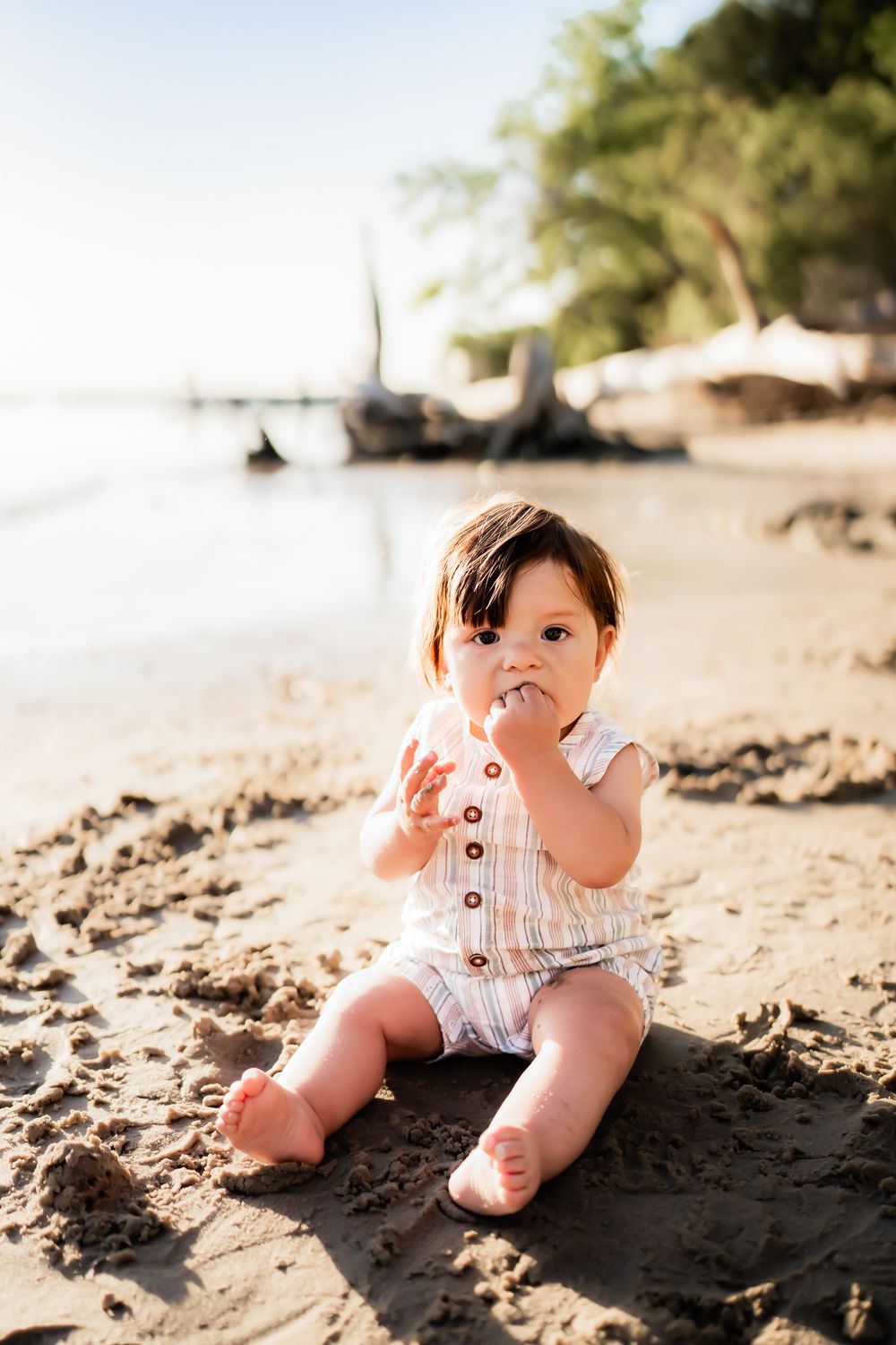 Baby wearing white romper sits on tropical beach sand during golden sunset.