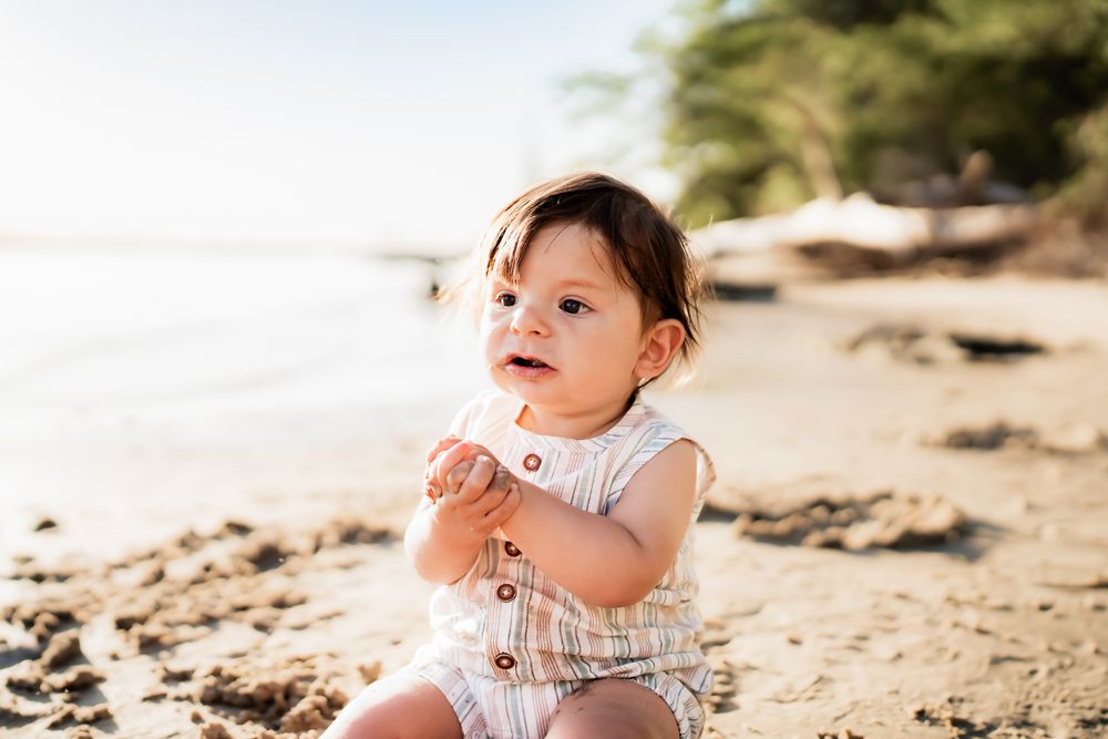 A small child in white clothing plays on a sunny tropical beach with sand and ocean in the background.