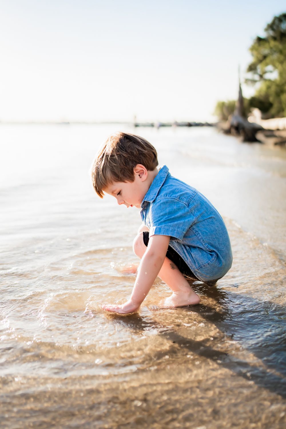 Young child in blue clothing crouches down to play in shallow water at a serene beach during golden hour.