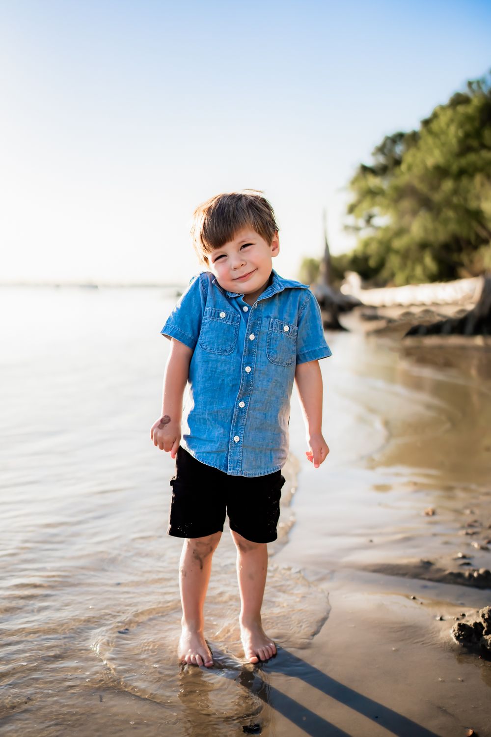 Child enjoying a sunny day at the beach wearing a blue shirt and black shorts while standing barefoot in shallow water.