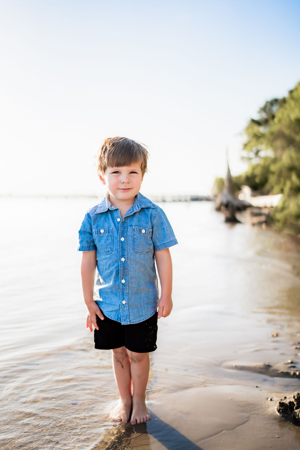 A young child wearing a blue denim shirt and dark shorts stands barefoot at the edge of a shoreline on a sunny day.