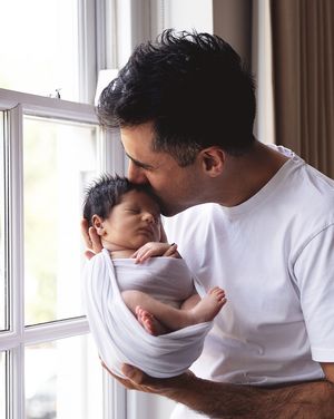 A tender moment by a window as an adult in a white t-shirt cradles and kisses a swaddled sleeping newborn baby. During a home newborn shoot