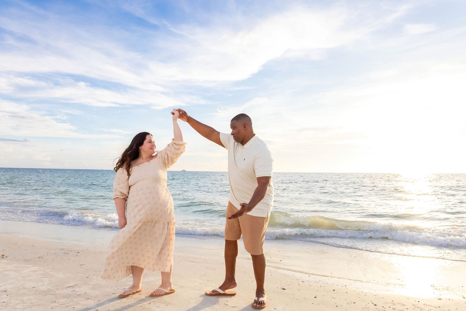 Couple in light colored outfits dancing playfully on beach at sunset.