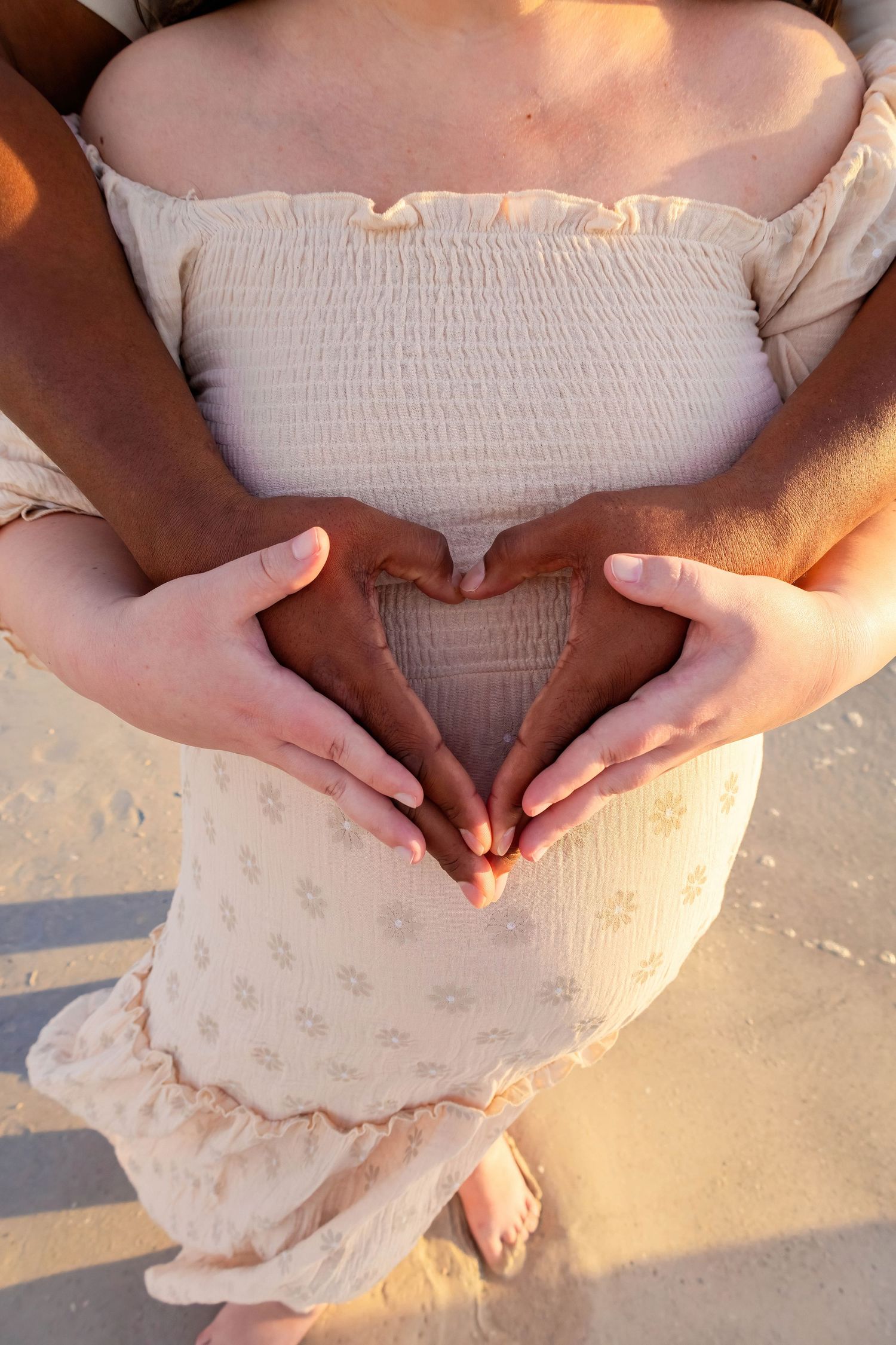 Intimate maternity photo showing hands forming heart shape on beach at sunset.