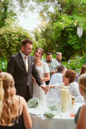 Couple interacting with guests at an elegant outdoor reception table with white linens and glowing decor at sunset.