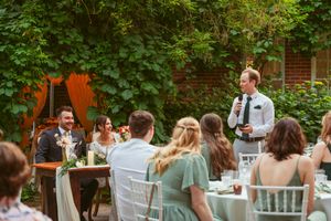 Guests enjoy an outdoor wedding reception in a garden setting with white chairs and tables under lush green foliage.