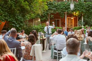 Wedding guests seated at outdoor reception while someone delivers a speech in front of a brick building.