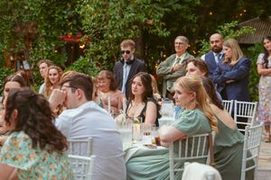 Wedding reception guests seated at white tables in an outdoor garden setting with natural greenery.