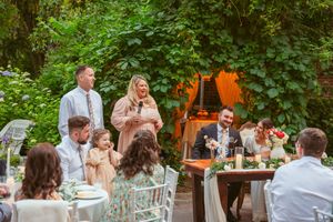 Outdoor wedding reception with guests dining at white tables surrounded by greenery.